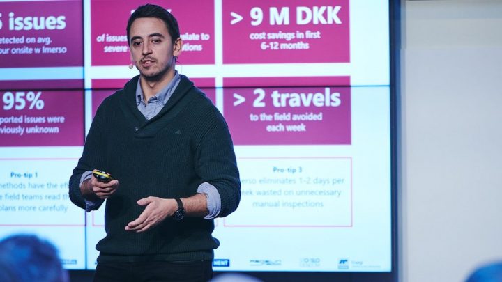A man presents at a conference, gesturing in front of a screen with stats on issues, cost savings, and travel reduction in fleet management.