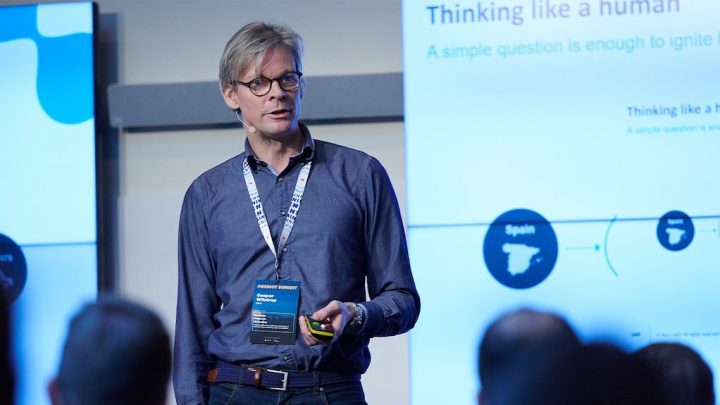 A man with glasses and a conference badge speaks on stage, remote in hand, as the audience listens to his talk on connected jobsite.