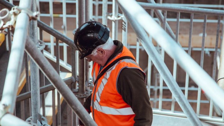 A person in a black hard hat and orange vest stands on scaffolding at a construction site, overseeing connected jobsite operations below.