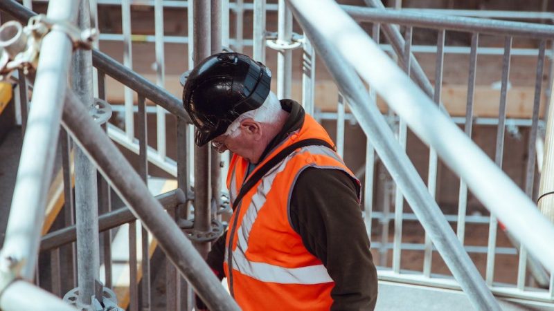 A person in a black hard hat and orange vest stands on scaffolding at a construction site, overseeing connected jobsite operations below.