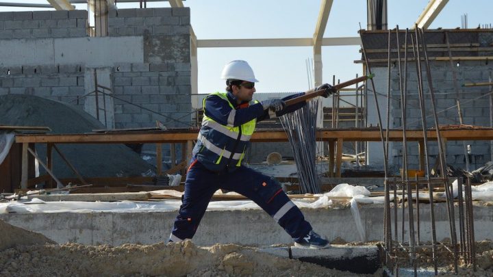 A construction worker in safety gear swings a sledgehammer at concrete on a connected jobsite with unfinished walls and metal rods.