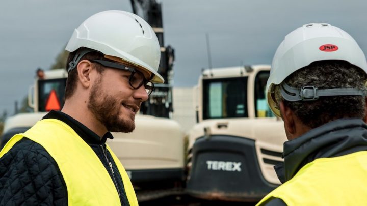 Two construction workers in safety gear talk at a worksite, with telematics-managed construction vehicles in the background.