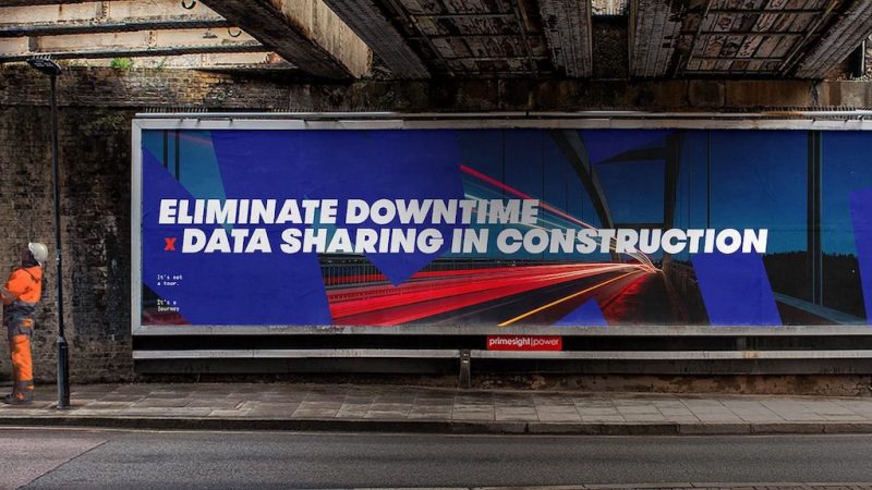 A construction worker in orange stands by a blue billboard under a bridge about data sharing to reduce downtime in construction.