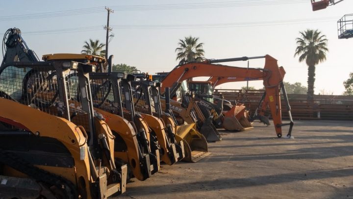 Yellow skid steers and excavators are lined up on a sunny lot with palm trees—fleet management in action.