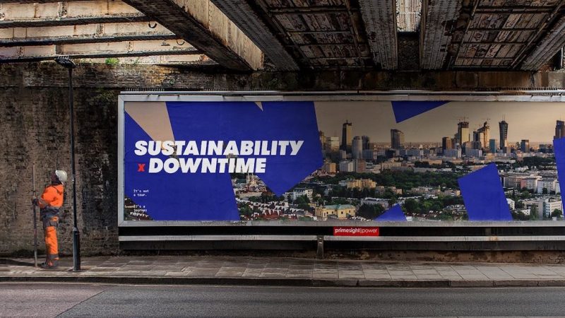 Worker in orange stands under a bridge by a "SUSTAINABILITY x DOWNTIME" billboard, suggesting connected jobsite themes.