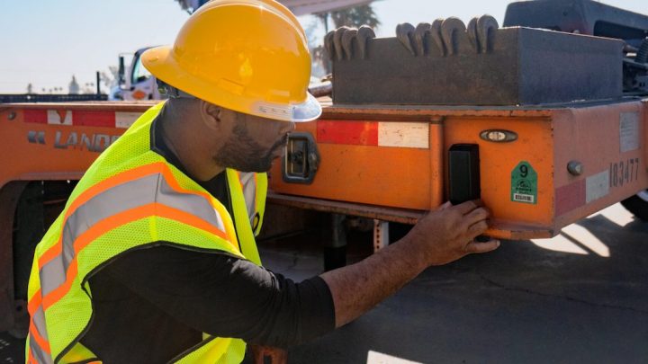 A construction worker in safety gear checks a large orange trailer with a telematics system at a job site, showing construction IoT.