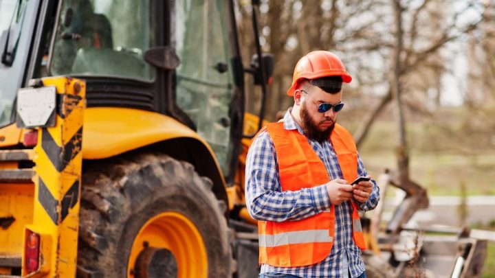 Construction worker in an orange vest and hard hat checks his phone by a yellow backhoe, possibly using telematics. Trees behind.