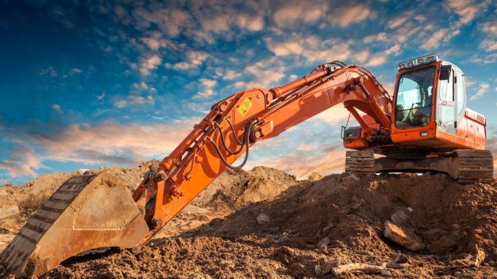 An orange excavator with telematics sits on a dirt mound at a construction site under a dramatic, partly cloudy sky.
