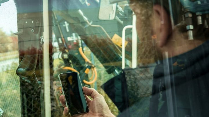 A person holds a smartphone, possibly using a fleet tracking app, looking at construction equipment through a glass window.