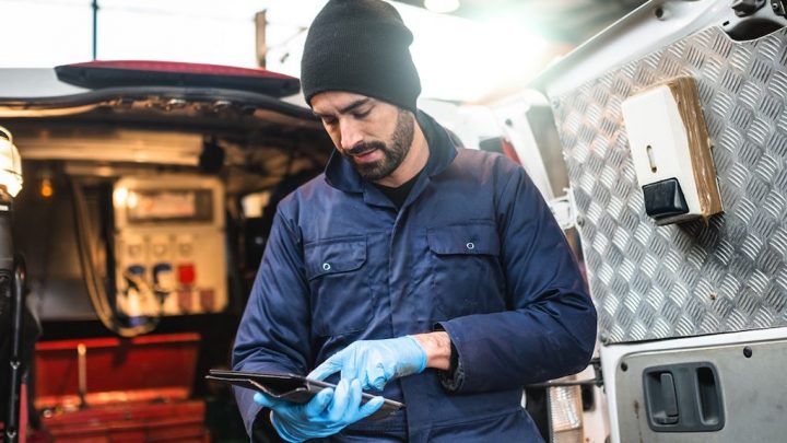 A mechanic in blue coveralls and gloves uses a tablet for asset tracking by an open van with tools, highlighting fleet management.