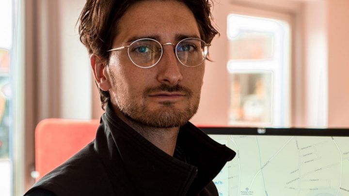 A man with wavy brown hair and glasses sits indoors; behind him, a screen shows equipment tracking software for construction.