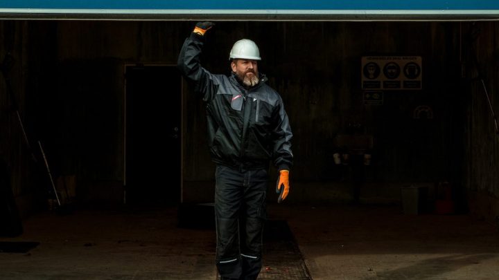 A man in a white hard hat and orange gloves holds open a garage door in a dim industrial space, suggesting a connected jobsite.
