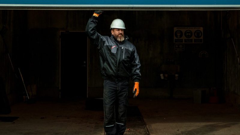 A man in a white hard hat and orange gloves holds open a garage door in a dim industrial space, suggesting a connected jobsite.