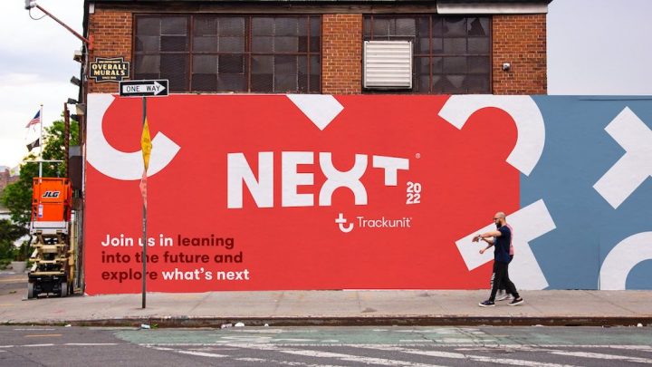 A person passes a colorful city mural reading "NEXT 2022 Trackunit," promoting the future of construction equipment tracking.
