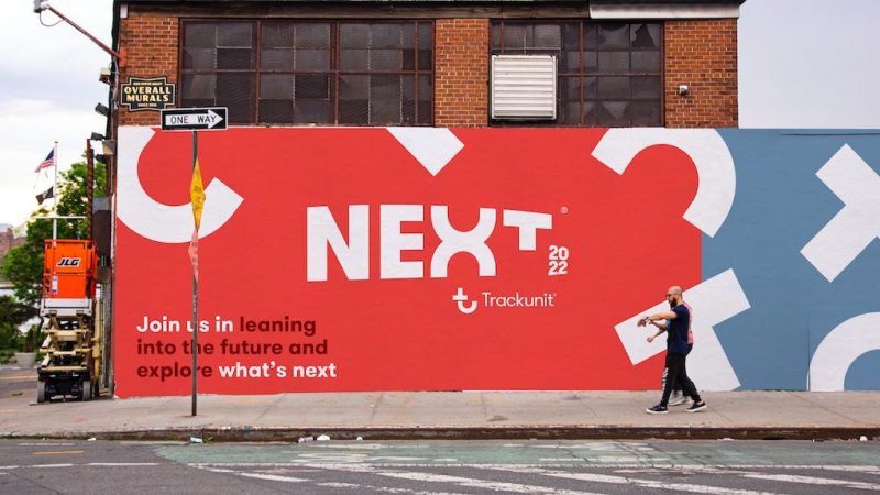A person passes a colorful city mural reading "NEXT 2022 Trackunit," promoting the future of construction equipment tracking.