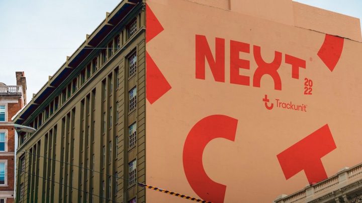 A peach-colored building with bold “NEXT 2022” text and the Trackunit logo is framed by older buildings.