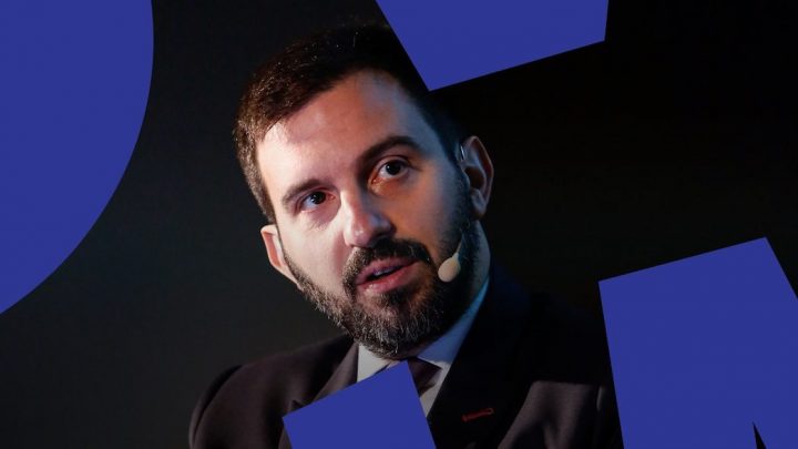 A bearded man in a suit with a headset mic speaks about inclusivity, dark background with blue shapes, related to connected jobsite.