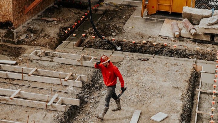 A construction worker in a red shirt and hard hat, award winner, walks through a connected jobsite with exposed foundations and materials.