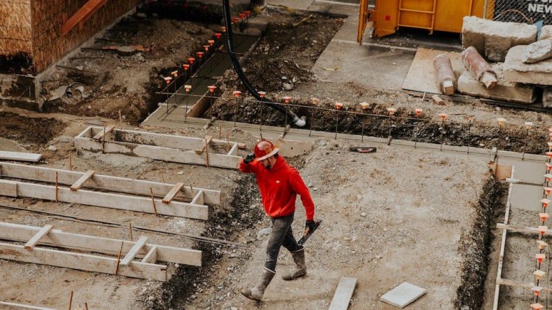 A construction worker in a red shirt and hard hat, award winner, walks through a connected jobsite with exposed foundations and materials.