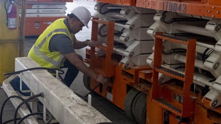 A worker in safety gear connects a charging cable to an orange scissor lift indoors, illustrating construction equipment tracking.