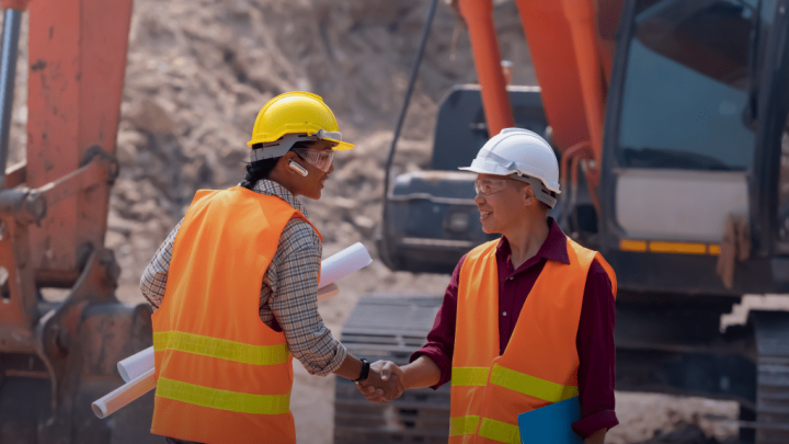 Two construction workers in safety gear shake hands at a connected jobsite with heavy machinery. One holds blueprints; both are smiling.