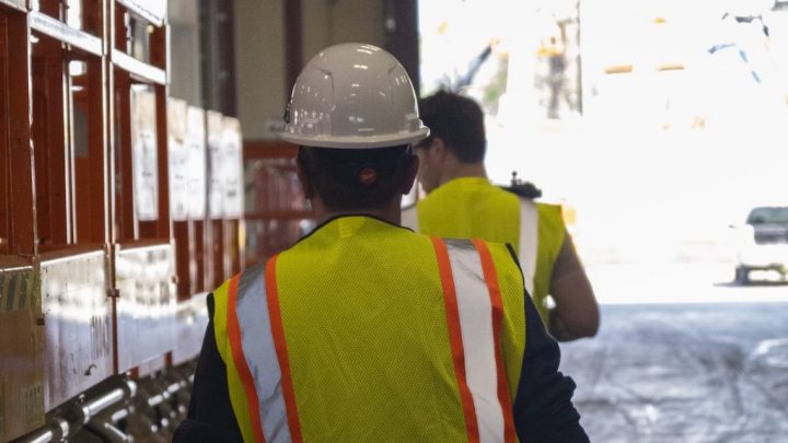 Two people in yellow safety vests and hard hats walk past orange equipment at an industrial site using fleet management.