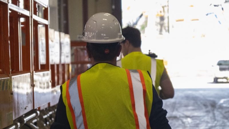 Two people in yellow safety vests and hard hats walk past orange equipment at an industrial site using fleet management.