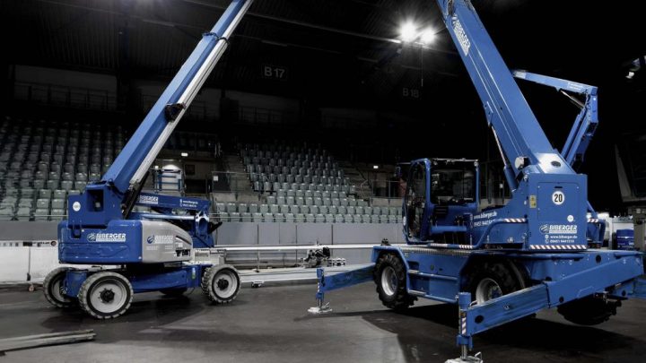 Two blue boom lifts are set up on the arena floor with tiered seating, likely for maintenance—an example of construction equipment tracking.
