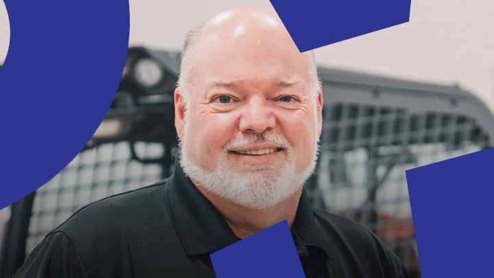 A smiling older man with a white beard stands in front of electrification equipment, with blue geometric shapes overlaid—connected jobsite.