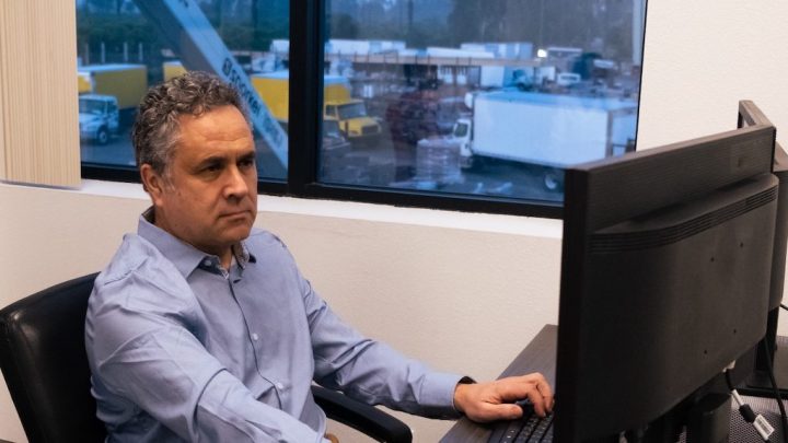 A man in a blue shirt uses tracking software at his desk, with trucks and equipment visible outside, highlighting fleet management.