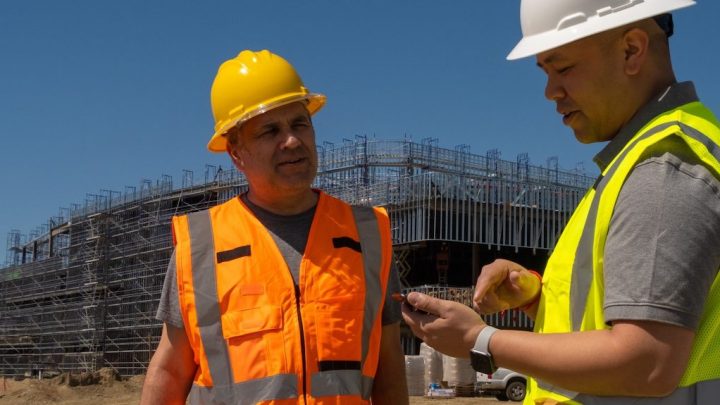 Two construction workers in safety gear discuss plans at a jobsite with scaffolding, using location tracking for construction efficiency.