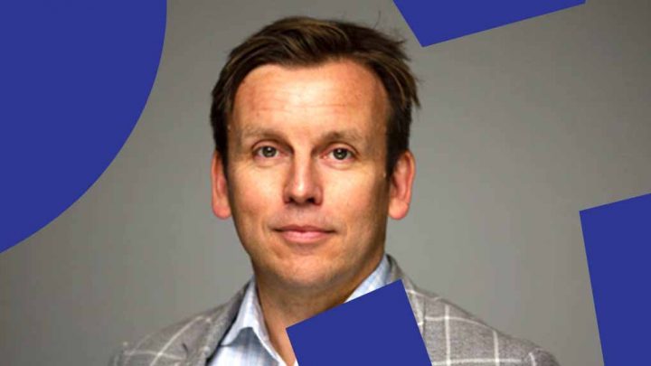 A man in a light blue shirt and gray plaid jacket poses before a plain background with blue geometric shapes, suggesting construction IoT.
