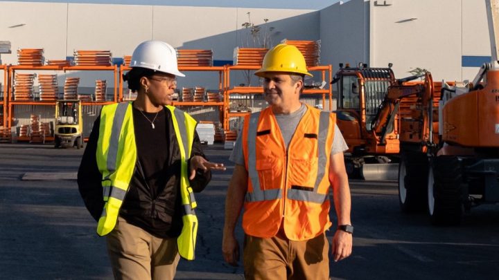 Two construction workers in safety gear walk and talk at an industrial site, highlighting connected jobsite technology.