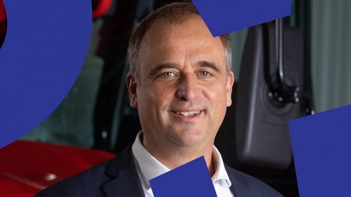 A man in a suit smiles at the camera in front of a red emission vehicle, with blue geometric shapes overlayed, suggesting fleet management.