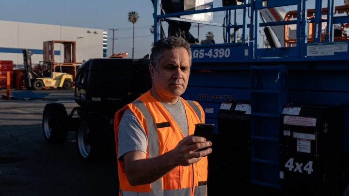 A man in a reflective orange vest uses his smartphone at a construction site with equipment in the background, showing telematics use.