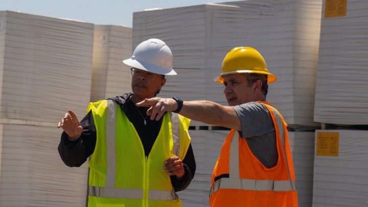 Two workers in safety gear discuss logistics by stacked pallets, using a GPS device for fleet management outdoors.