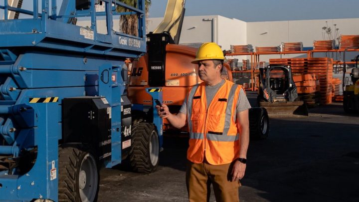 A man in a yellow hard hat and orange vest operates a blue scissor lift by remote in an equipment yard, highlighting fleet management.