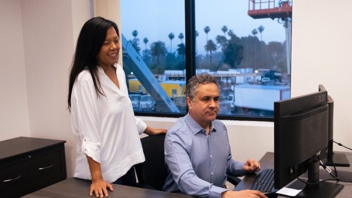 A woman stands beside a man at a desk with a computer in an office, discussing fleet management as trees and buildings show outside.