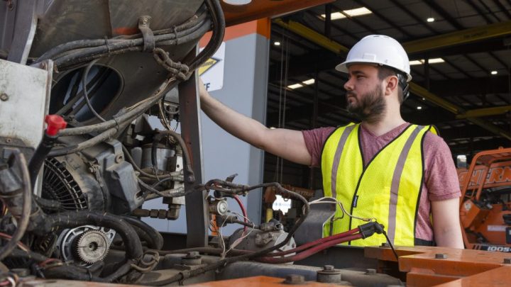A man in a hard hat and safety vest inspects machinery and telematics systems at an industrial site, highlighting telematics.