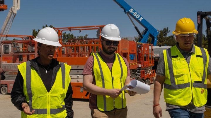 Three workers in yellow vests and hard hats walk at a construction site, one holds blueprints; equipment and crane highlight construction IoT.