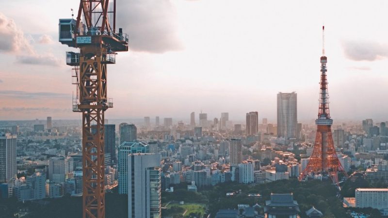 A sunset cityscape with a tall orange crane on the left and Tokyo Tower on the right, highlighting construction equipment tracking.