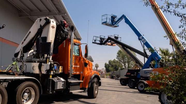 An orange utility truck and blue boom lifts are parked by an industrial building, with workers in baskets—fleet management equipment.