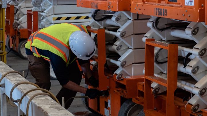 A construction worker inspects machinery in a warehouse with orange and white equipment, emphasizing construction fleet management.