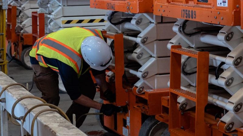A construction worker inspects machinery in a warehouse with orange and white equipment, emphasizing construction fleet management.