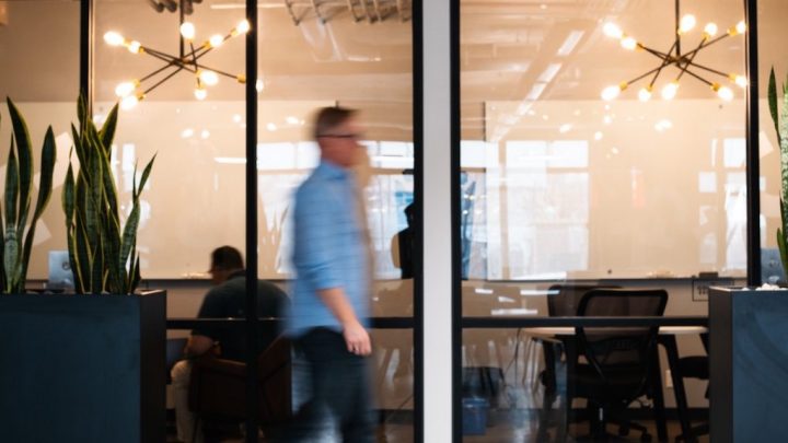 A blurred man in a blue shirt walks past a glass-walled office where people collaborate on AI projects for connected jobsite solutions.
