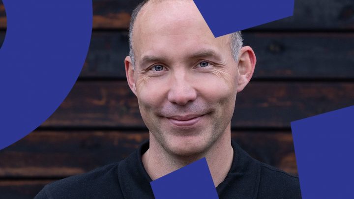 A smiling man with a shaved head stands before a dark wooden wall and blue shapes, reflecting calm on a connected jobsite.