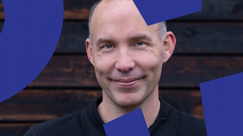 A smiling man with a shaved head stands before a dark wooden wall and blue shapes, reflecting calm on a connected jobsite.