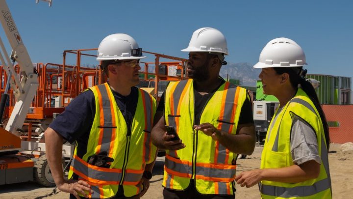 Three construction workers in safety gear talk at a job site with equipment and fencing behind them, highlighting fleet management.