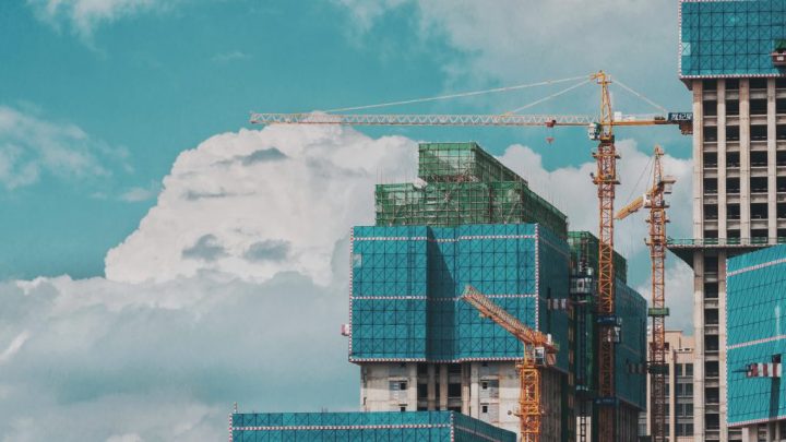 High-rise buildings under construction with blue netting and yellow cranes, showcasing a connected jobsite beneath a bright sky.