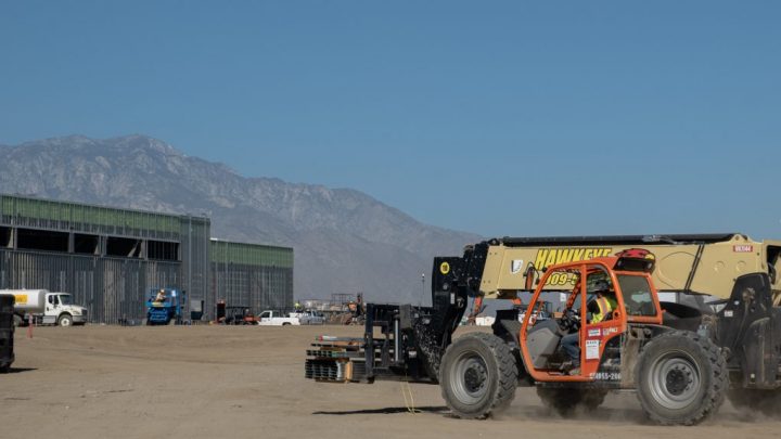 A forklift with telematics moves materials on a construction site with buildings and mountains in the background under a blue sky.
